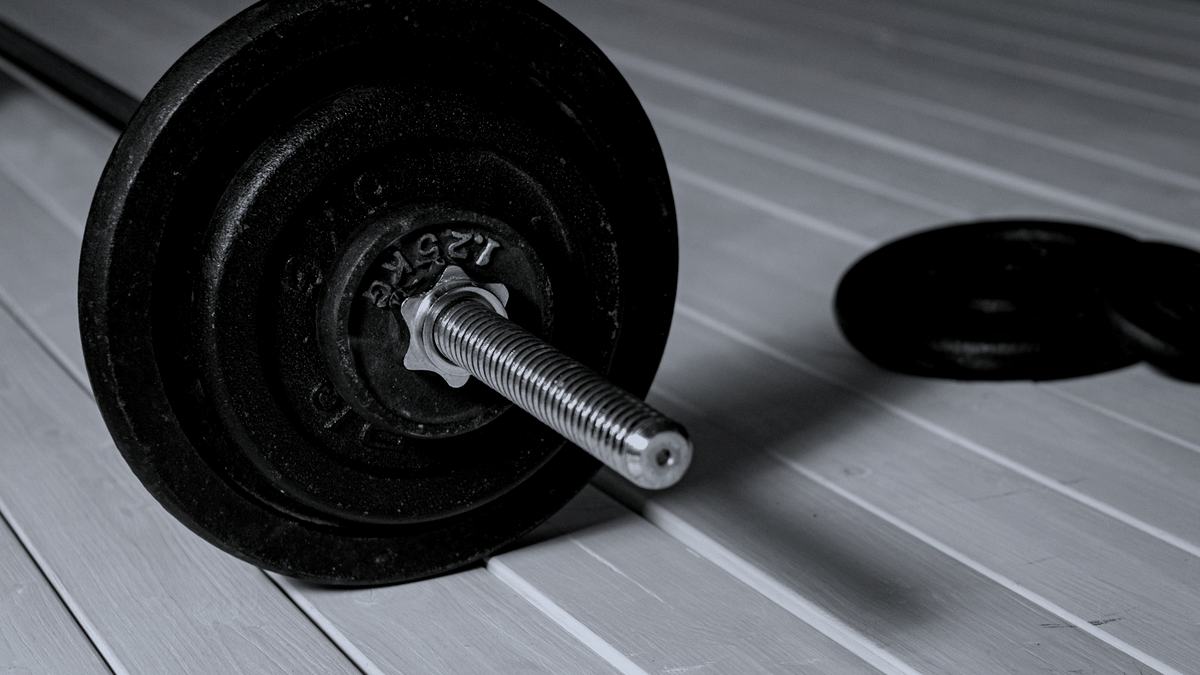 A loaded barbell lying on a pale wooden floor with spare plates behind it, in near-monochrome.