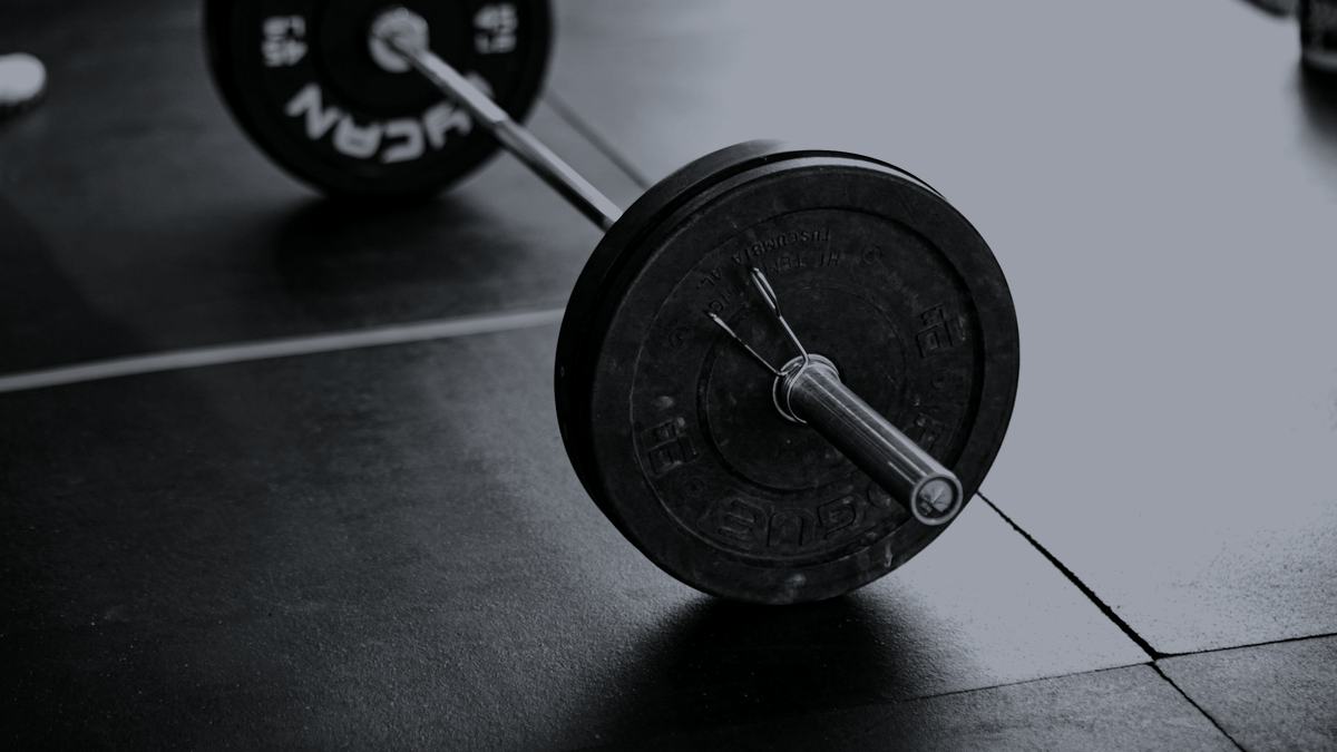 A bumper-plated barbell lying on a rubber gym floor, shot low and close in near-monochrome.